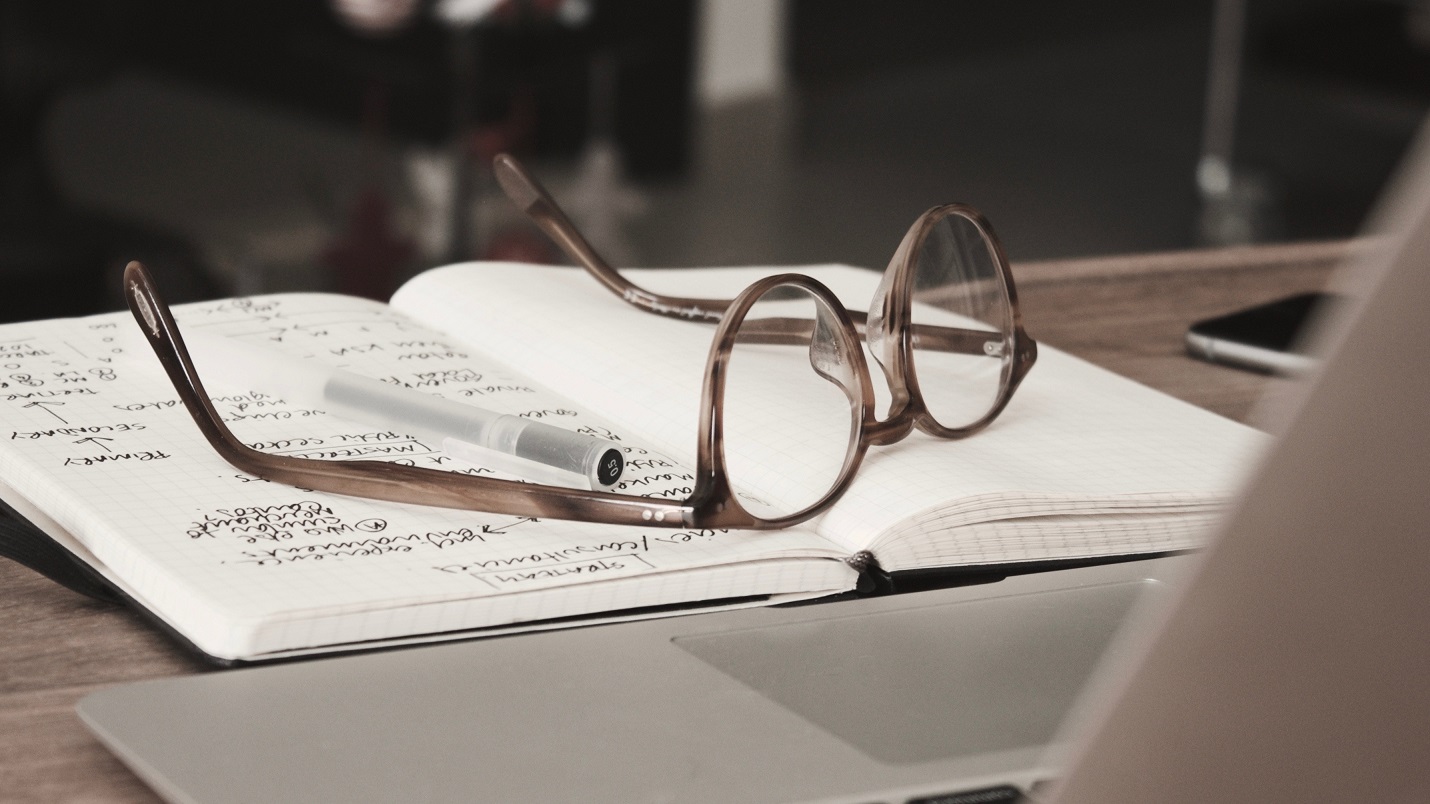 open notebook with pen and glasses on it in front of a laptop