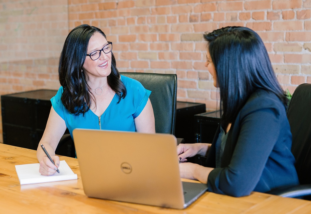 two women talking at a conference table; one has a laptop, the other is taking notes