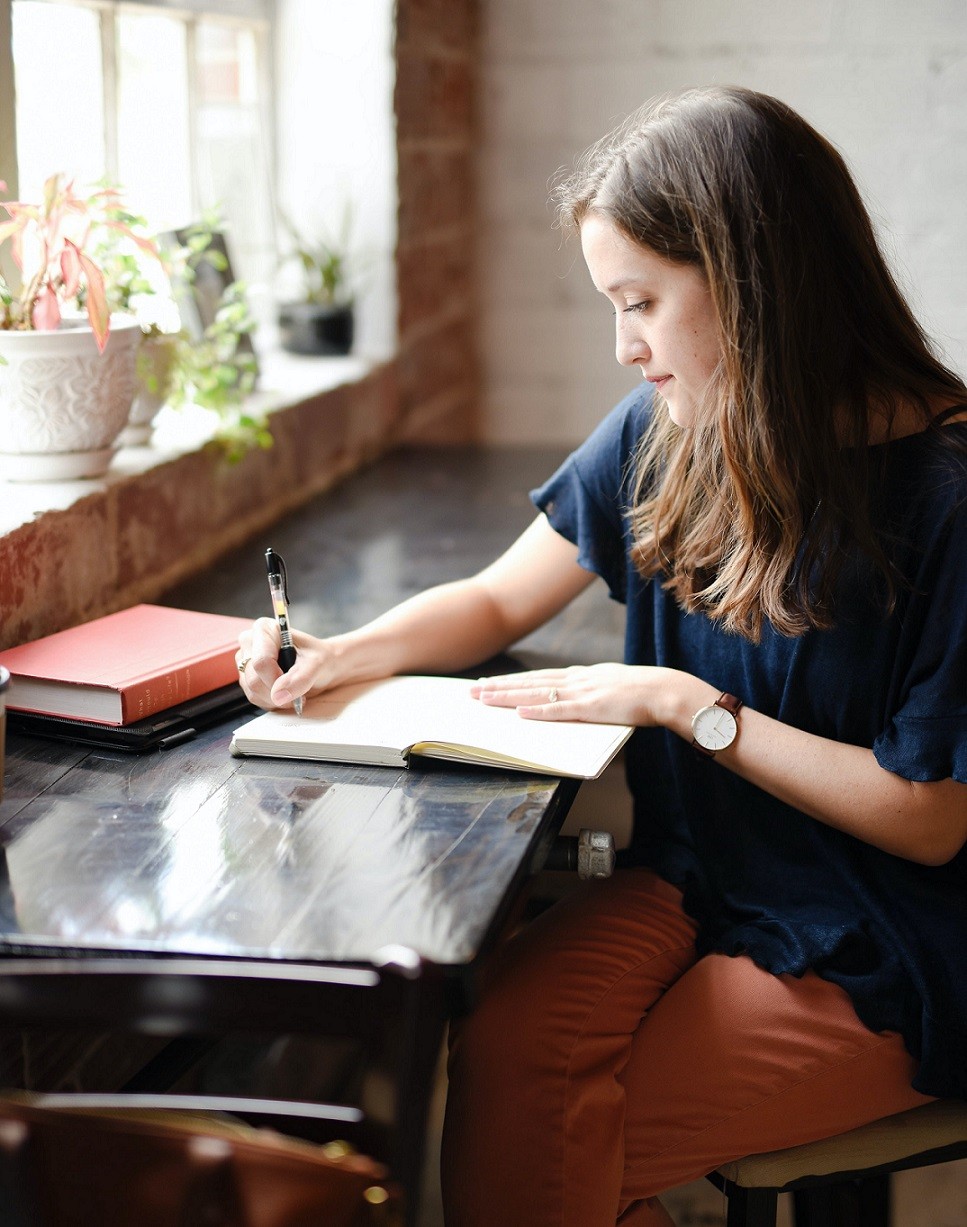 female writing in journal sitting next to window