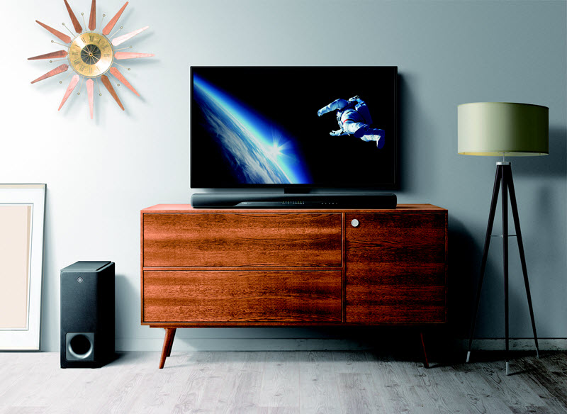 YAS207 Entertainment area of a living room setup with a sun shaped clock on wall, a wood credenza with a flat screen TV and sound bar on top and a subwoofer and a floor lamp on rug on floor.