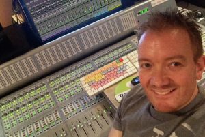 Man in his 30's dressed in t-shirt and smiling for the camera while sitting in front of a sound studio audio console.