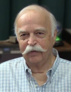 An older Caucasion man with white hair and handlebar moustache in open necked checked shirt smiling for camera.