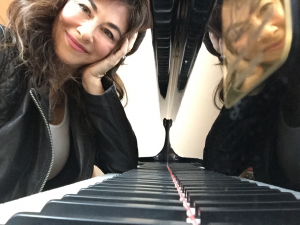 Female songwriter, Shelly Peiken, resting her elbow on the piano keyboard while smiling into camera.