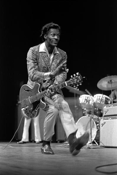 chuck berry watermarked African American man in white pants and sports jacket with an electric guitar duckwalking across a stage with other musicians and drums in background.