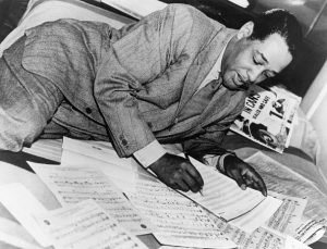 duke ellington watermarked Tall African American man in a windowpane patterned suit and tie laying on his side on a bed while writing out musical notation.