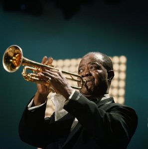 louis armstrong watermarked Middle-aged African American man in a suit onstage playing a trumpet.