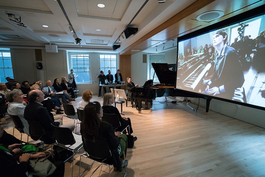 Pianos without borders body image 1 A group of people watch a presentation by a pianist in a large room with a projector.