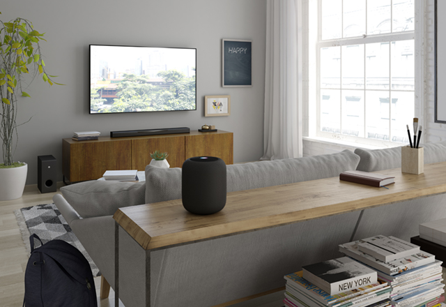 homepod in room Modern living room with couch facing flat screen on wall in background and a table in foreground with small cylindrical speaker type of electronics on table.