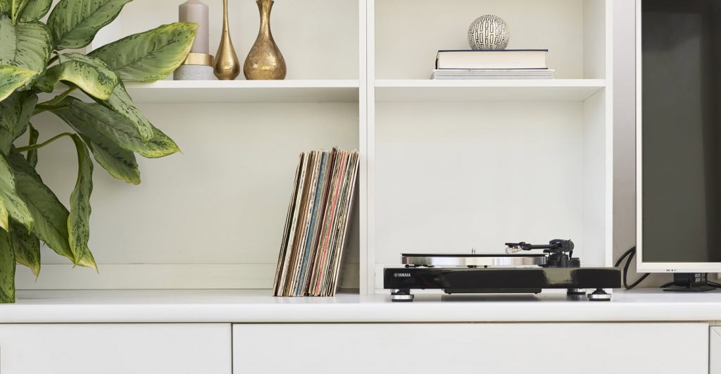 Turntable on a shelf next to a flat screen and a row of vinyl albums in their dust covers, plus other decorative items on the shelves above.