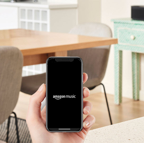 In a room where there is a table and chairs in background, you see a hand holding a smartphone with the screen showing the Amazon Music logo.