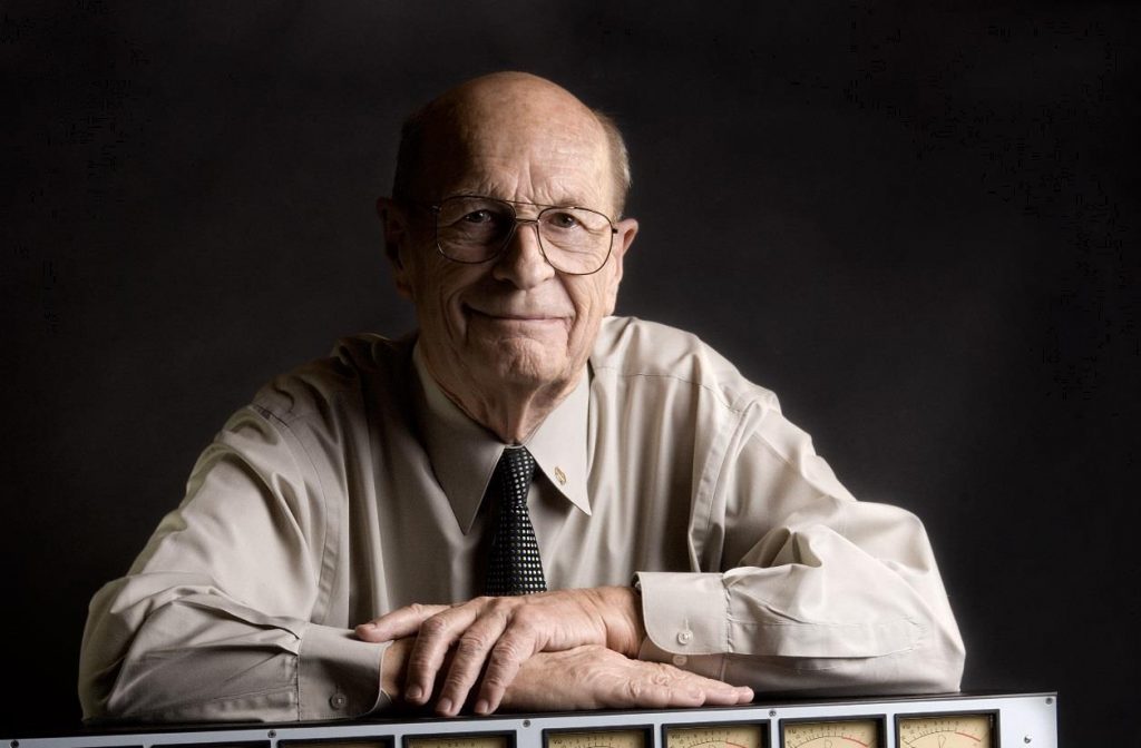 rupert neve connection body image 1 Older man in shirt and tie with his forearms resting on an older version of a sound board.
