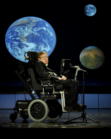 stephen hawking Man in a suit slumped down in an electric wheelchair with a computer screen in front of him and images of Earth and other planets projected in the background.