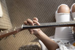 storia body image 1400 Looking over the shoulder of a guitar player, a closeup of their hand fingering the strings on the neck. You can see that they are sitting in a living room, wearing casual clothes.