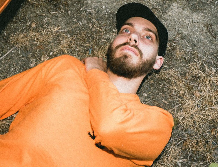 Bearded young man in long sleeved t-shirt and billed cap laying on ground staring upwards.