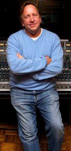 Middle-aged man in jeans and sweater casually leaning against a sound board.