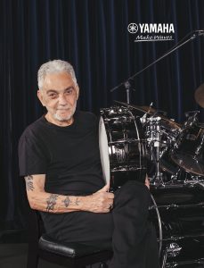 Tattooed man with white hair holding snare drum, sitting next to drum kit in front of purple curtains.