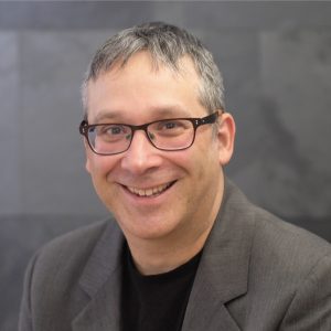 Gary Marcus Portrait of man in suit jacket smiling at camera in front of grey studio backdrop.