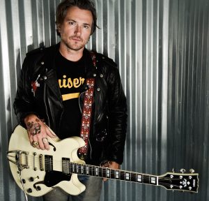 BW-new-body-image-1-1200.jpg Man in t-shirt and jacket with an electric guitar slung over his shoulders.