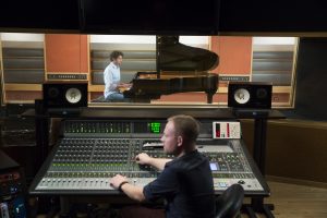 Man behind mixing desk recording a pianist in a sound booth.