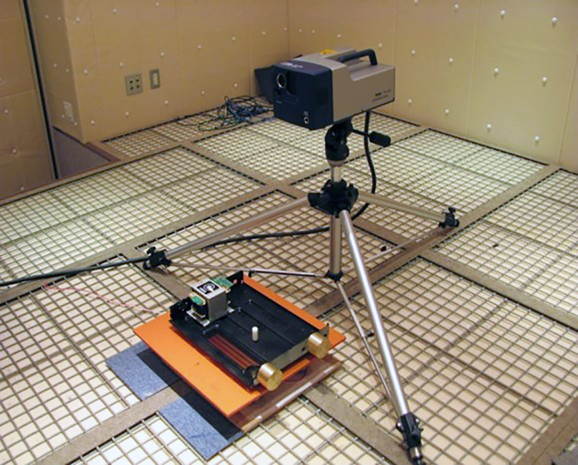 003-Laser-Doppler-VibrometerII Tripod holding a small rectangular unit in a room with a grid-tracked floor. There is a small electronic unit on a pad that is on grid track in front of the unit on a tripod.