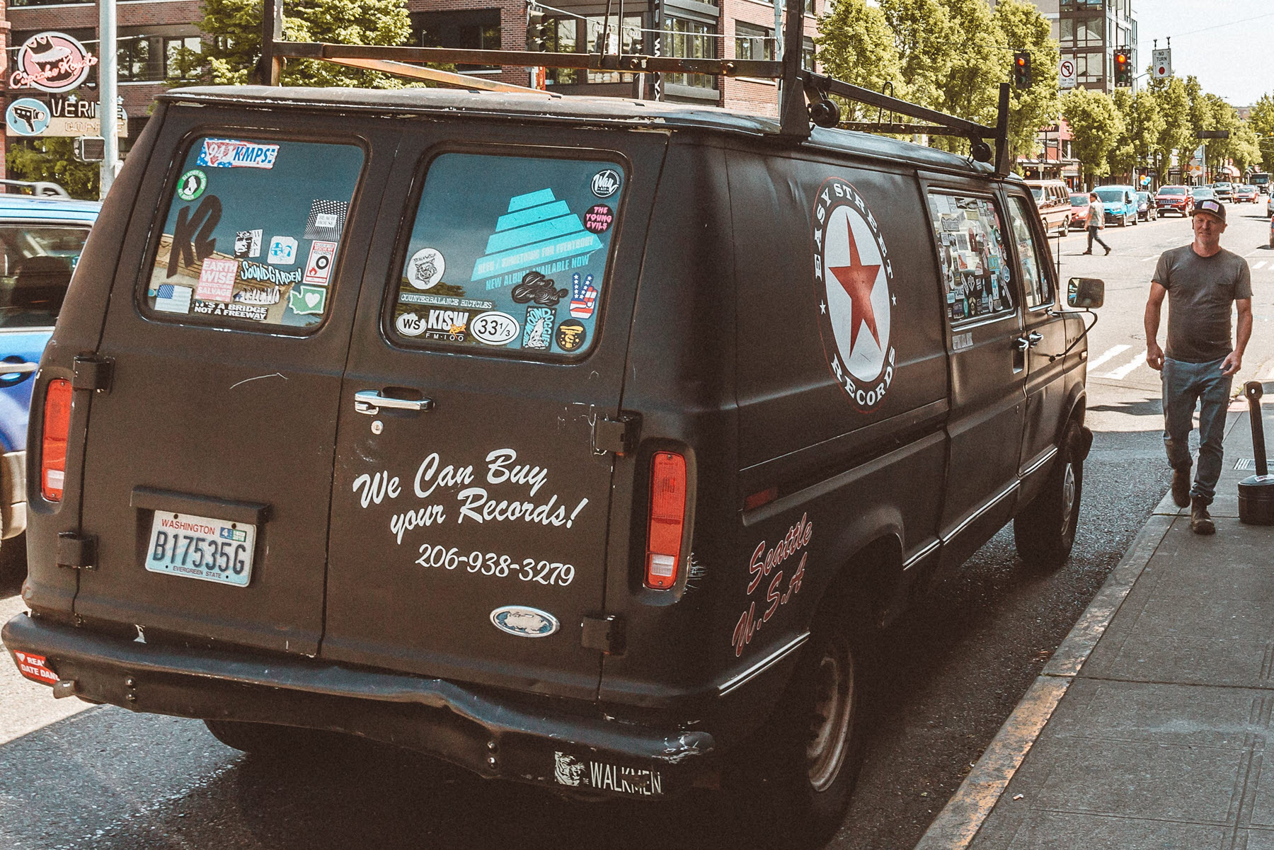 Easy_Street_Van-cropresize. View of a well-used cargo van with stickers for record store and "We can buy your records" and their phone number. It is parked on a street and man walking towards it.