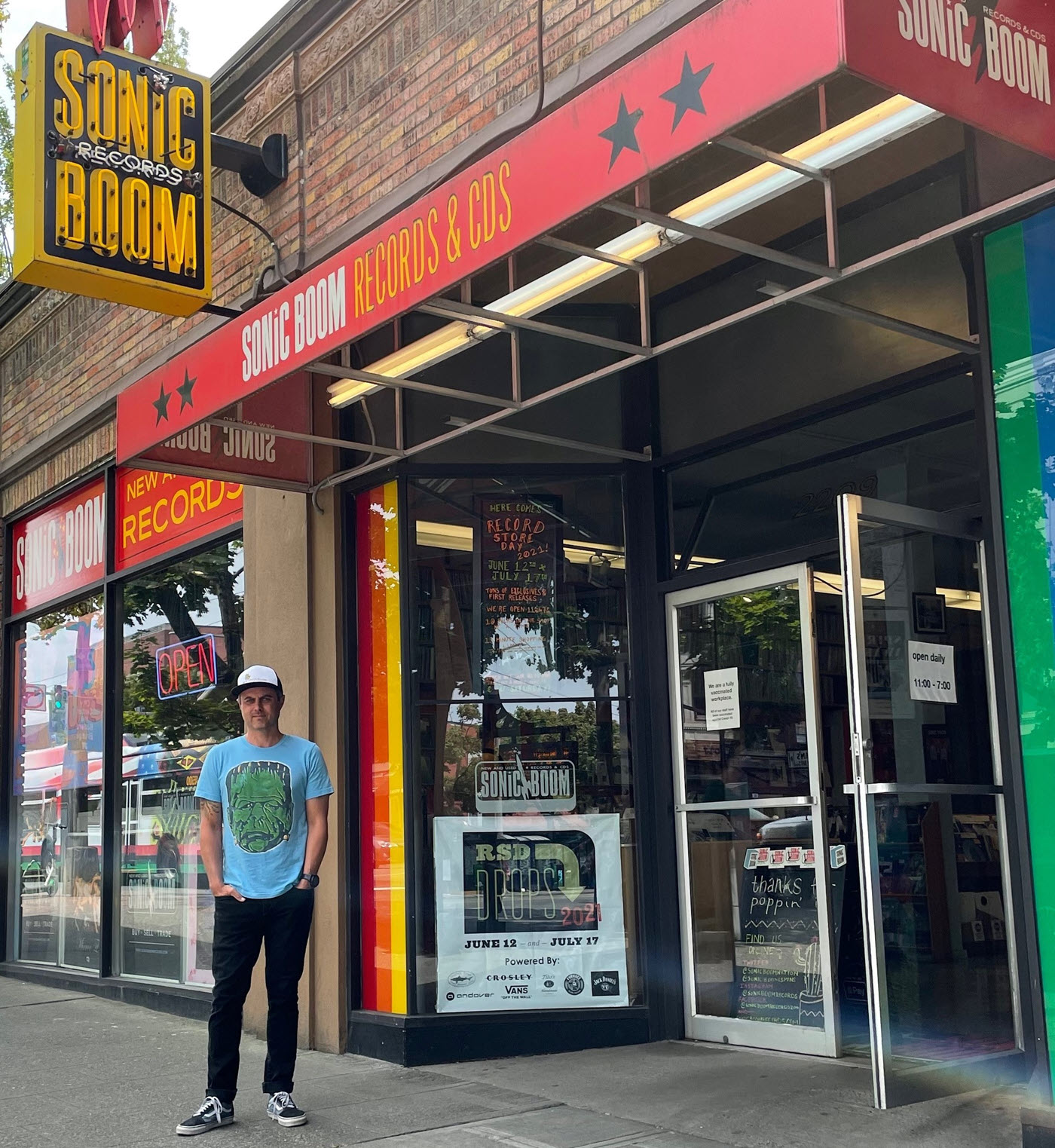 Sonic_Boom_Pitts-cropresize Man in graphic t-shirt, jeans and cap standing in front of urban record store with signage indicating it is "Sonic Boom Records & CDs".