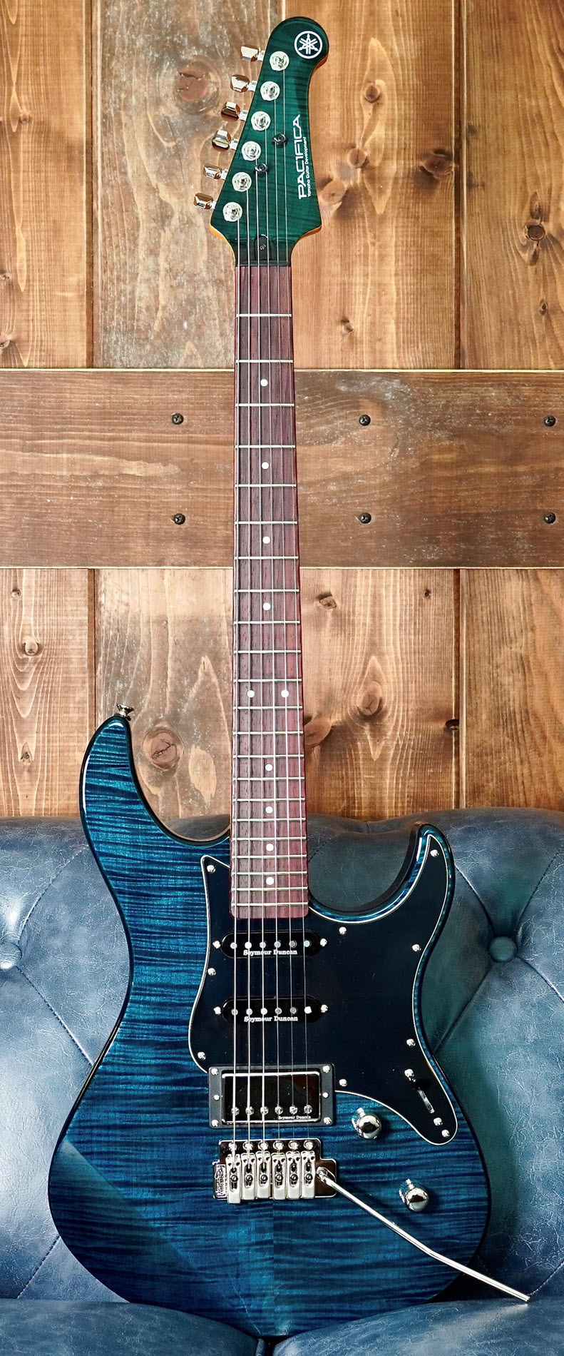 Pacifica-crop-1900 Beautiful blue electric guitar standing upright on a blue couch with a wood wall behind it.