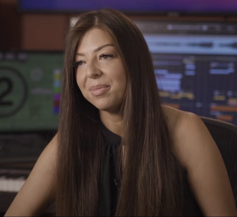 Young woman with long dark hair seated in front of a digital sound studio smiling for camera.