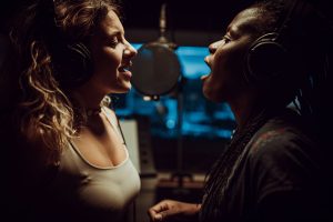 Two female singers on either side of a microphone.