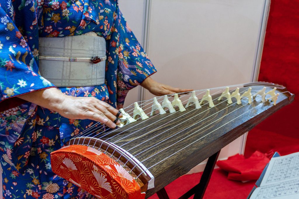 Koto-resize Closeup of a woman in kimono with a long rectangular stringed instrument across her lap. She is playing the strings with her right hand.