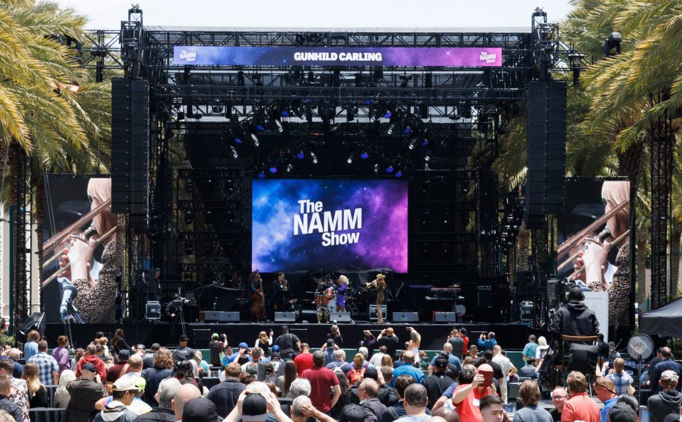 Main-Stage1-crop Crowd seen from behind facing an outdoor stage where a band is playing and you can see a screen behind the with "The NAMM Show" projected on it.