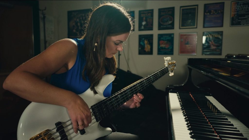 Woman playing an electric guitar while seated adjacent to a grand piano.