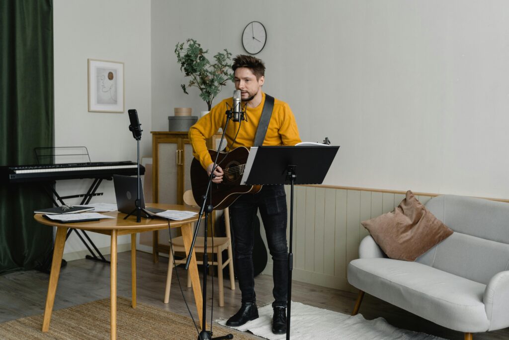 A man playing an acoustic guitar in a room with a music stand in front of him, a curtain behind him, a small throw rug beneath his feet, plus a cushioned seat, a small cabinet in the corner, plus a wood table and an electronic keyboard on a stand.