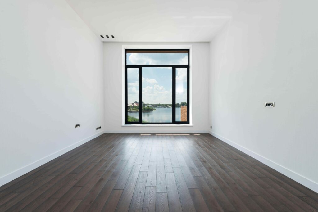 A room with parallel sheetrock walls, wood flooring, and a large window at one end.